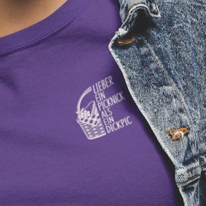 Könnte beinhalten: Ein violettes T-Shirt mit einer weißen Grafik eines Picknickkorbs und dem Text "LIEBER EIN PICKNICK ALS EIN DICKPIC" auf der linken Brustseite, getragen unter einer blauen Acid-Wash-Jeansjacke mit sichtbarem Kragen und Metallknöpfen.
