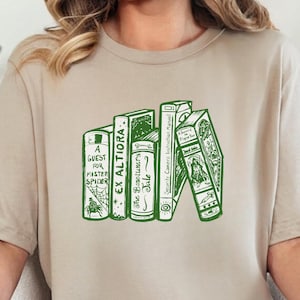 Könnte beinhalten: Beiges T-Shirt mit einem grünen Aufdruck von gestapelten Büchern, darunter Titel wie "A Guest for Master Spyder" und "Ex Altiora." Das Shirt hat eine lockere Passform und wird mit blauen Jeans getragen. Ein goldenes Armband ist ebenfalls zu sehen.