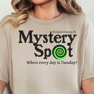 Könnte beinhalten: Ein beiges T-Shirt mit den schwarzen Worten "Mystery Spot" und einem grünen Spiraldesign in der Mitte des Wortes "Spot". Der Text "Where every day is Tuesday!" steht darunter. Das T-Shirt wird von einer Person getragen.