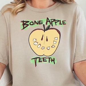 Könnte beinhalten: Ein beiges T-Shirt mit den grünen Worten "Bone Apple Teeth", das eine gelbe Apfelgrafik mit Zähnen umrahmt. Das T-Shirt wird mit einer blauen Jeans kombiniert.