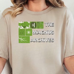 May include: A beige t-shirt with the text "THE MAGNUS ARCHIVES" in a white font. The text is above a green and white graphic. The person is wearing blue jeans.