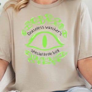 Puede incluir: Camiseta beige con un gr&aacute;fico verde ne&oacute;n. El dise&ntilde;o dice "Ceaseless watcher's special little boy" y muestra un ojo. La persona lleva vaqueros azules.