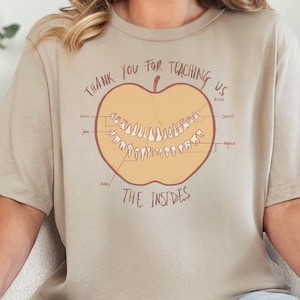 Könnte beinhalten: Beiges T-Shirt mit einer Grafik eines halbierten Apfels, der Zähne zeigt und mit "The Insides" beschriftet ist. Der Text darüber lautet "Thank You For Teaching Us". Das Shirt wird mit blauen Jeans getragen.