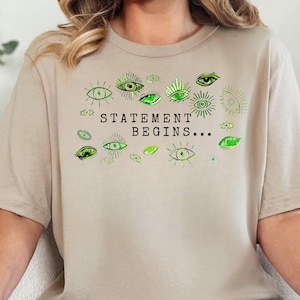 Könnte beinhalten: Beiges T-Shirt mit dem Text "STATEMENT BEGINS..." in Schwarz und Grün. Das Shirt hat grüne Augengrafiken. Die Person trägt eine blaue Jeans.