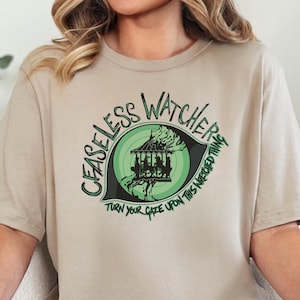 Könnte beinhalten: Beiges T-Shirt mit dem Text "CEASELESS WATCHER" in Grün und Schwarz, sowie einer Karussell-Grafik in einem grünen Auge. Der Text "TURN YOUR GAZE UPON THIS NEEDED THING" ist ebenfalls vorhanden. Das T-Shirt wird getragen.