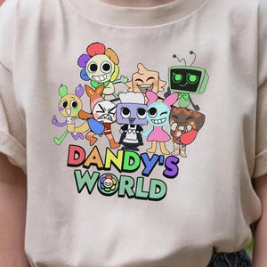 Może przedstawiać: Beżowa koszulka z kolorową grafiką postaci z kresk&oacute;wek i napisem "DANDY'S WORLD" w wielokolorowej czcionce. Projekt zawiera r&oacute;żne fantazyjne stworzenia i zabawną estetykę.