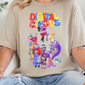 May include: A beige t-shirt featuring the text "THE AMAZING DIGITAL CIRCUS" above a colorful cartoon illustration of various characters. The shirt is worn by a person in blue jeans.
