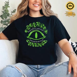Puede incluir: Camiseta negra con un dise&ntilde;o de ojo verde y el texto "Ceaseless watcher's special little boy". El dise&ntilde;o presenta un ojo estilizado con una pupila verde, rodeado de formas de ojos verdes m&aacute;s peque&ntilde;as. La camiseta es de cuello redondo cl&aacute;sico.