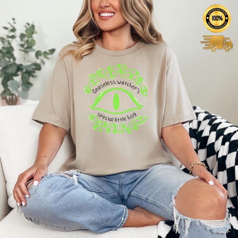 Puede incluir: Camiseta beige con un gr&aacute;fico verde ne&oacute;n. El dise&ntilde;o dice "Ceaseless watcher's special little boy" y muestra un ojo. La persona lleva vaqueros azules.
