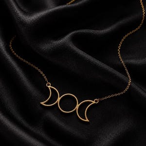 May include: A gold-toned necklace featuring a central pendant shaped as a triple moon symbol, consisting of two crescent moons flanking a full circle. The pendant is attached to a delicate, thin gold-toned chain with a simple link design.