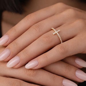 May include: A gold-coloured ring in the shape of a cross, set with small, clear gemstones. The ring is worn on a finger, with the hand and other fingers visible. The nails are painted a light pink colour.