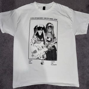 Op de afbeelding: Wit t-shirt met een zwart-wit afbeelding van twee vrouwen met gitaren, met de tekst "STRAWBERRY SWITCHBLADE" erboven. Onder de afbeelding staan de woorden "I HATE THE TREES" en "I HATE THE FLOWERS".