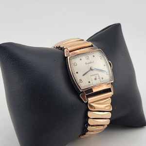 May include: A vintage gold-toned wristwatch with a rectangular face and a stretch band. The watch face is white with black numerals and hands, and the brand name "Corteluart" is visible. The watch is displayed on a black cushion.
