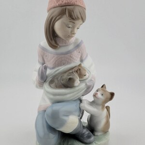May include: A porcelain figurine of a girl in a winter hat and sweater, holding a sack with two kittens. A third kitten stands nearby, reaching up. The figurine is painted in soft pastel colours, with a green base.