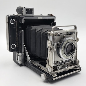 May include: A vintage black and silver folding press camera with a black accordion bellows, a metal lens board, and a circular lens assembly. The side features the text Kalart Synchronized Range Finder, and the top includes a viewfinder attachment.