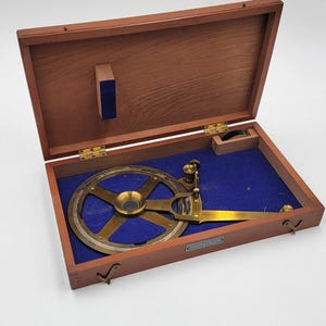 Puede incluir: Un antiguo instrumento de topografía de latón y madera en una caja de madera abierta. El instrumento presenta un componente circular de latón con una abertura central y una escala graduada. La caja tiene un forro de fieltro azul y bisagras de latón.