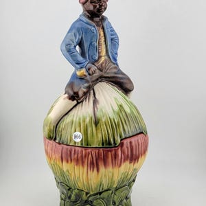 May include: A ceramic figurine of a person in a blue jacket and red hat, perched atop a colorful, onion-shaped container. The container has green, yellow, and red hues. The number "966" is on a small white sticker on the container.