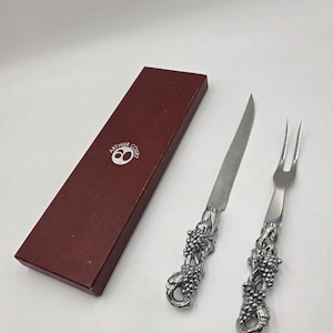 May include: A silver cheese knife and fork set with grape cluster handles, next to a maroon box. The knife has a long, thin blade, and the fork has two prongs. The box has the text "ARTHUR COURT" and a logo.