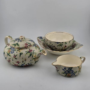 May include: A ceramic tea set featuring a teapot, a teacup with a saucer, and a creamer. Each piece has a cream-colored base with a fine black grid pattern and a colorful floral design in blue, red, and yellow, finished with gold-toned rims.