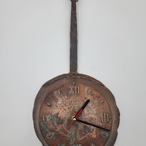 May include: A rustic, copper-toned wall clock with a handle. The clock face features Roman numerals and decorative fish motifs. The clock hands are black with a red second hand. The clock is mounted on a white wall.