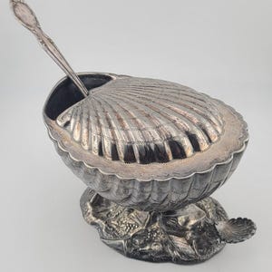 Puede incluir: Un plato para servir plateado en forma de concha con una cuchara a juego. El plato se asienta sobre una base decorativa con un diseño de concha. La tapa está abierta, revelando el interior oscuro. La cuchara descansa dentro del plato.