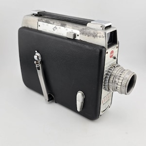 Puede incluir: Cámara de cine Kodak 8mm vintage con una cubierta de cuero negro y detalles metálicos plateados. La cámara tiene una lente con marcas de medición y un asa en la parte superior. El logotipo de Kodak es visible. Una pieza clásica de equipo de fotografía de película.