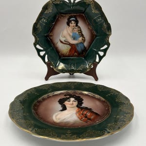 Puede incluir: Dos platos decorativos de porcelana con bordes verde oscuro y patrones de filigrana dorada. El plato superior es hexagonal con bordes recortados y muestra a una mujer abrazando a un niño. El plato redondo inferior muestra el retrato de una mujer.
