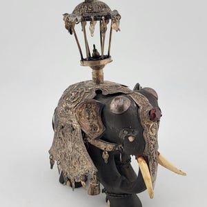 Puede incluir: Una detallada y decorativa figura de elefante con adornos plateados y una estructura en forma de cúpula en la espalda. El elefante es negro con colmillos de marfil y detalles rojos. Pequeñas campanas cuelgan de los adornos plateados.