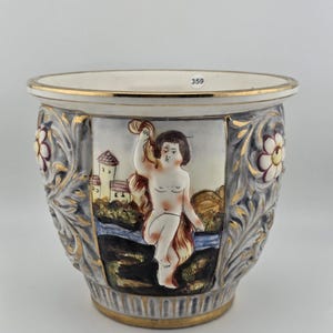 Puede incluir: Una maceta de cerámica con un relieve central de una figura sentada cerca de edificios, flanqueada por motivos florales en gris y dorado. El borde y la base están adornados con oro, y una pequeña etiqueta blanca con el número 359 es visible en el borde interior.