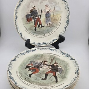 Może przedstawiać: Stos ceramicznych talerzy z napisem LES GRANDES MANŒUVRES oraz szczegółowymi ilustracjami żołnierzy w mundurach. Krawędzie ozdobione są niebieskim wzorem kwiatowym, a sceny na środku przedstawiają historyczne postacie wojskowe.