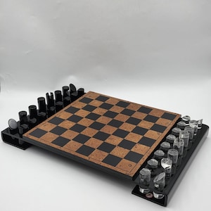May include: A chess set featuring a square board with a checkered pattern of brown cork and black squares. The board is framed in black and includes two integrated side trays holding chess pieces. One side holds solid black pieces, while the other side holds clear, transparent pieces.