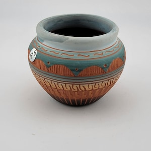 May include: A small, round ceramic pot featuring a light blue rim and a terracotta-coloured body. The exterior is decorated with teal bands, carved semi-circular patterns, and vertical lines. A small white sticker with the number 515 is attached.