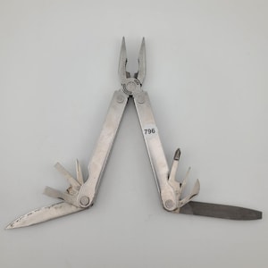 May include: A silver-coloured stainless steel multi-tool with pliers in the centre and various folded implements extending from the handles. A small white circular sticker with the black text 796 is affixed to one of the metal handles.