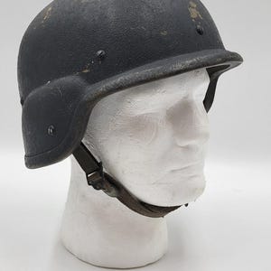 以下が含まれることがあります： 黒色のミリタリースタイルのヘルメット。表面はテクスチャ加工され、イヤープロテクターとあご紐が付いています。白い発泡スチロール製の頭部に展示されています。ヘルメットはわずかに使用感があります。