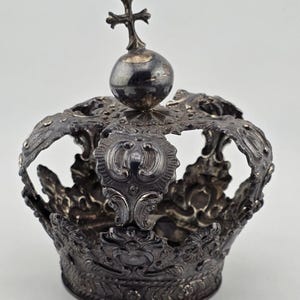 May include: An antique silver crown with elaborate detailing, including a cross atop a sphere. The crown's design incorporates intricate patterns and floral motifs. The object is likely a decorative piece, possibly for display or ceremonial use.