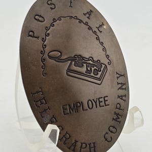 Puede incluir: Insignia ovalada de bronce de empleado de Postal Telegraph Company. La insignia presenta el nombre de la empresa y la palabra «EMPLOYEE» en el centro, con una ilustración de una máquina telegráfica. La insignia se muestra sobre un soporte transparente.