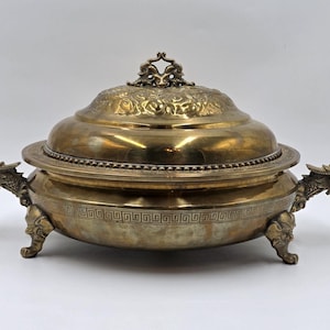 Puede incluir: Una fuente de servir vintage en tono latón con tapa, con asas ornamentadas en forma de cabeza de dragón y detalles decorativos. La fuente tiene forma redonda, soportada por tres patas de garra y un detallado patrón floral en la tapa.