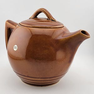 May include: A brown ceramic teapot with a lid and handle. The teapot has a rounded body, a spout, and a handle on the side. The lid has a small handle on top. The teapot is a glossy brown color and has a number 95 sticker on the side.