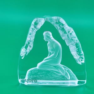 May include: A clear glass sculpture featuring a frosted figure of a person kneeling on a rock, framed by an arched, textured glass structure. The base includes the etched text Nybro Crystal Sweden, showcasing a detailed, translucent design.