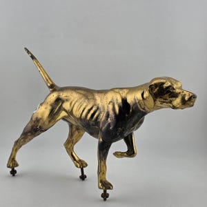 May include: A golden-colored dog statue with a dark patina, possibly bronze. The dog is in a poised stance, with one front leg raised. The statue has a detailed, realistic design and is mounted on small, dark metal feet.