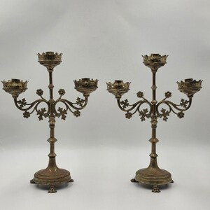 May include: Two antique brass candelabras. Each candelabra has a central column with three arms, each holding a candle cup. The arms are adorned with floral and leaf designs. The base is a four-footed design.