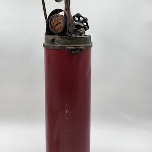 May include: A vintage, cylindrical fire extinguisher with a deep red body and a rusty, metallic top. The top features a pressure gauge, a handle, and a valve. The extinguisher is likely made of metal and shows signs of age and use.