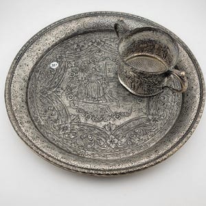 Puede incluir: Un juego de taza y platillo plateado antiguo. La taza tiene dos asas y se apoya en una bandeja redonda a juego. Ambas piezas presentan intrincados grabados florales y figurativos. La bandeja tiene un diseño de borde detallado. El juego es probablemente vintage.