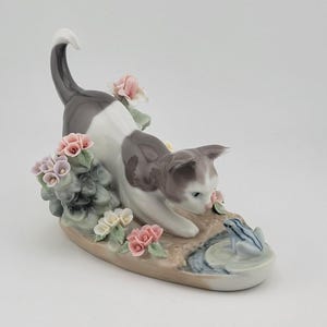 Puede incluir: Figura de porcelana de un gato gris y blanco jugando con una rana azul y blanca. El gato está rodeado de flores rosas y blancas y vegetación, sobre una base marrón claro.