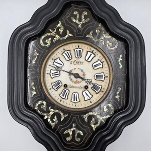 May include: A black wall clock with a scalloped frame and mother-of-pearl inlay in floral patterns. The circular face features Roman numerals, a central floral wreath, and the text E CHERY and Chateaumeillant printed in black ink.
