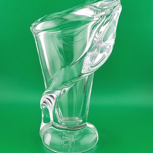 Puede incluir: Un florero de vidrio transparente con un diseño escultural y asimétrico, que presenta una abertura ancha y acampanada junto a una base gruesa y redondeada. El cuerpo del florero muestra formas de vidrio fluidas que crean una silueta dinámica.