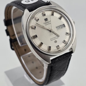Op de afbeelding: Een vintage Tissot Swiss Automatic Seastar horloge met een zilveren wijzerplaat en een zwarte leren band. De wijzerplaat toont de dag en de datum. Het horloge heeft een roestvrijstalen kast en een zwarte leren band.