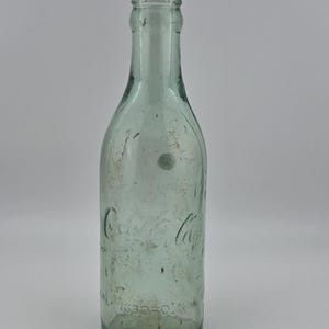 May include: A vintage, light green glass Coca-Cola bottle with a narrow neck and a flared rim. The glass features embossed lettering that reads Coca-Cola and Roseboro, N.C. The surface of the bottle shows signs of wear, including minor scuffs.