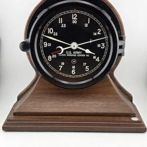 May include: A vintage U.S. Army clock with a black face and white numerals. The clock has a dark wooden base and a black bezel. The text "U.S. ARMY CLOCK. MESSAGE CENTER. M2" is printed on the face. The clock is approximately 20 cm in height.