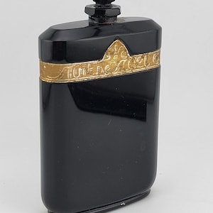 Może przedstawiać: Czarny, prostokątny flakon perfum ze złotą opaską z napisem "Fume de Noel". Butelka ma czarny, fasetowany korek. Projekt jest inspirowany stylem Art Deco.